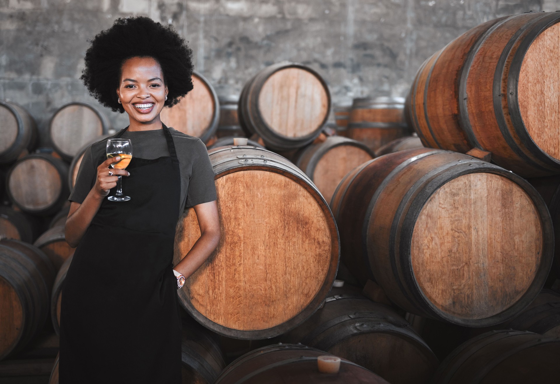 Portrait of a young woman winemaker standing with a glass with wooden barrel of red wine in a winery cellar or distillery. Entrepreneur or business owner working for startup success business success